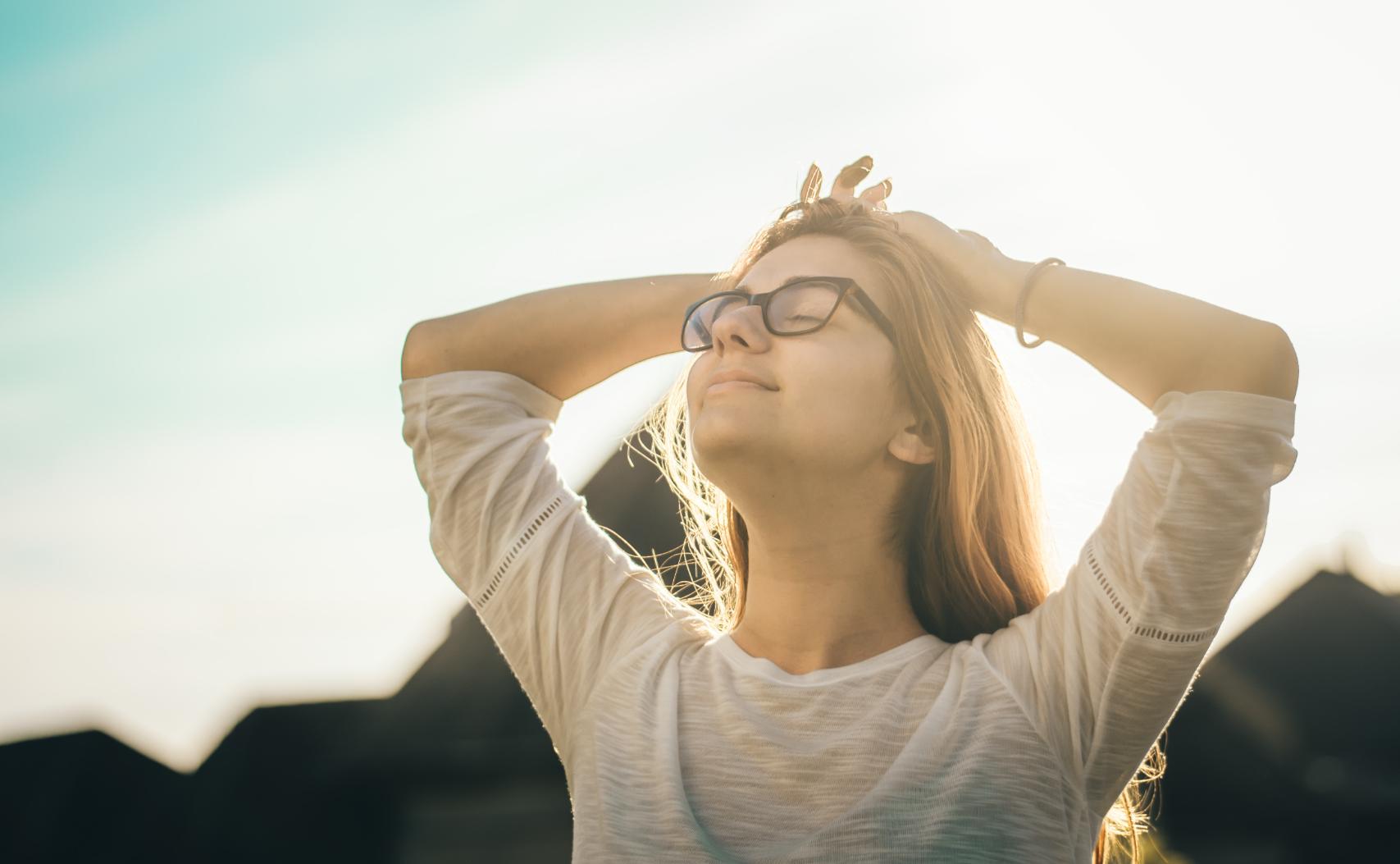 Mujer con anteojos con los brazos sobre su cabeza y los ojos cerrados tomando el sol en pos de tranquilidad y felicidad